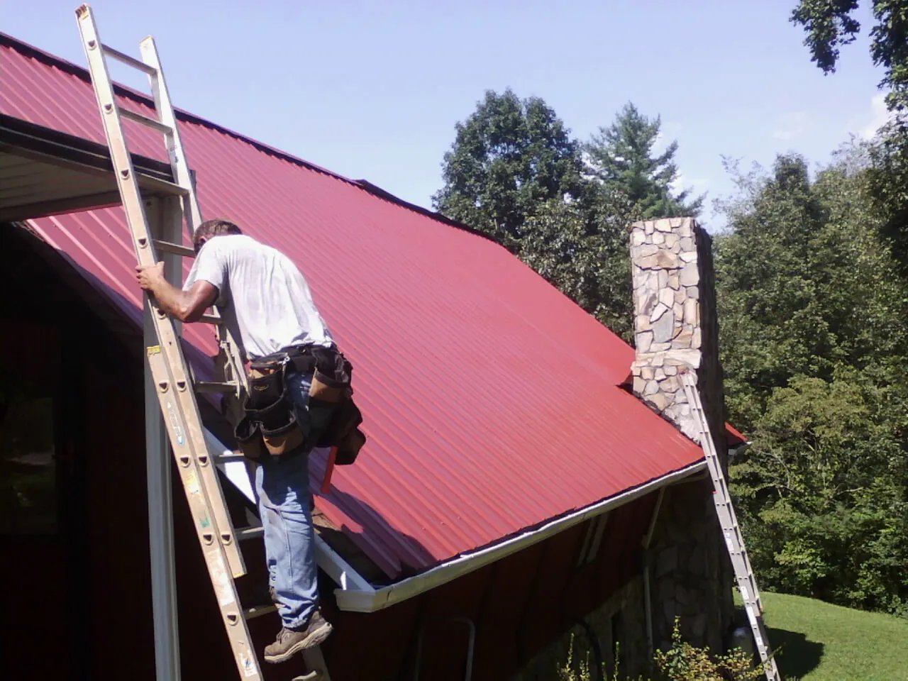 Roofer on a ladder beside a red metal roof and stone chimney — Soffit & Fascia Repair by Roofing Brothers in Macclenny